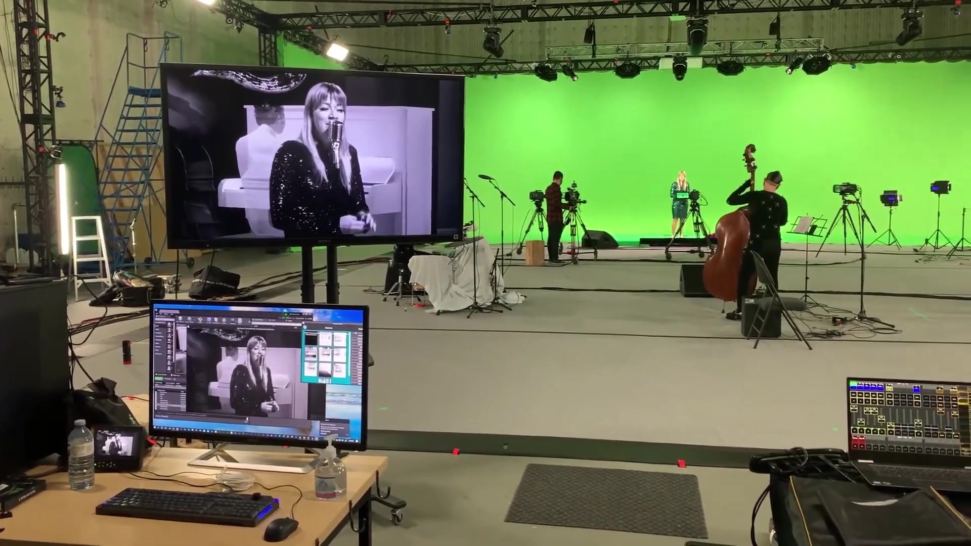 Behind the scenes at Animatrik's studio during the live broadcast: Jill Barber performing live on the green-screen stage (right of frame) while her band plays alongside her — the bass player visible at her side. The monitor at left shows the live composite output, with Jill rendered onto the virtual Palomar stage in real time. The operator's console at the bottom of frame runs the live show.